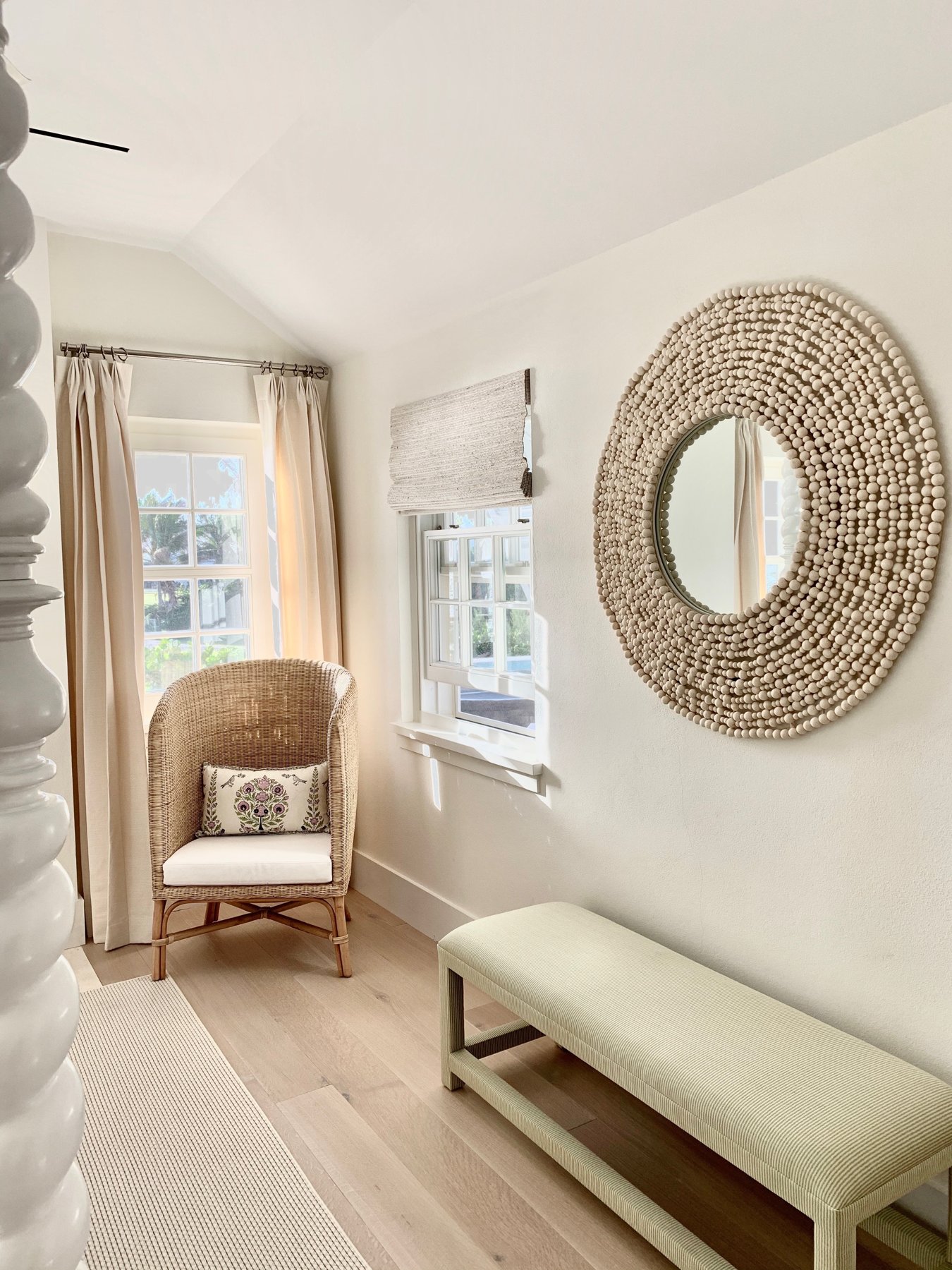Rattan chair beside a window with a round woven mirror and a pale bench