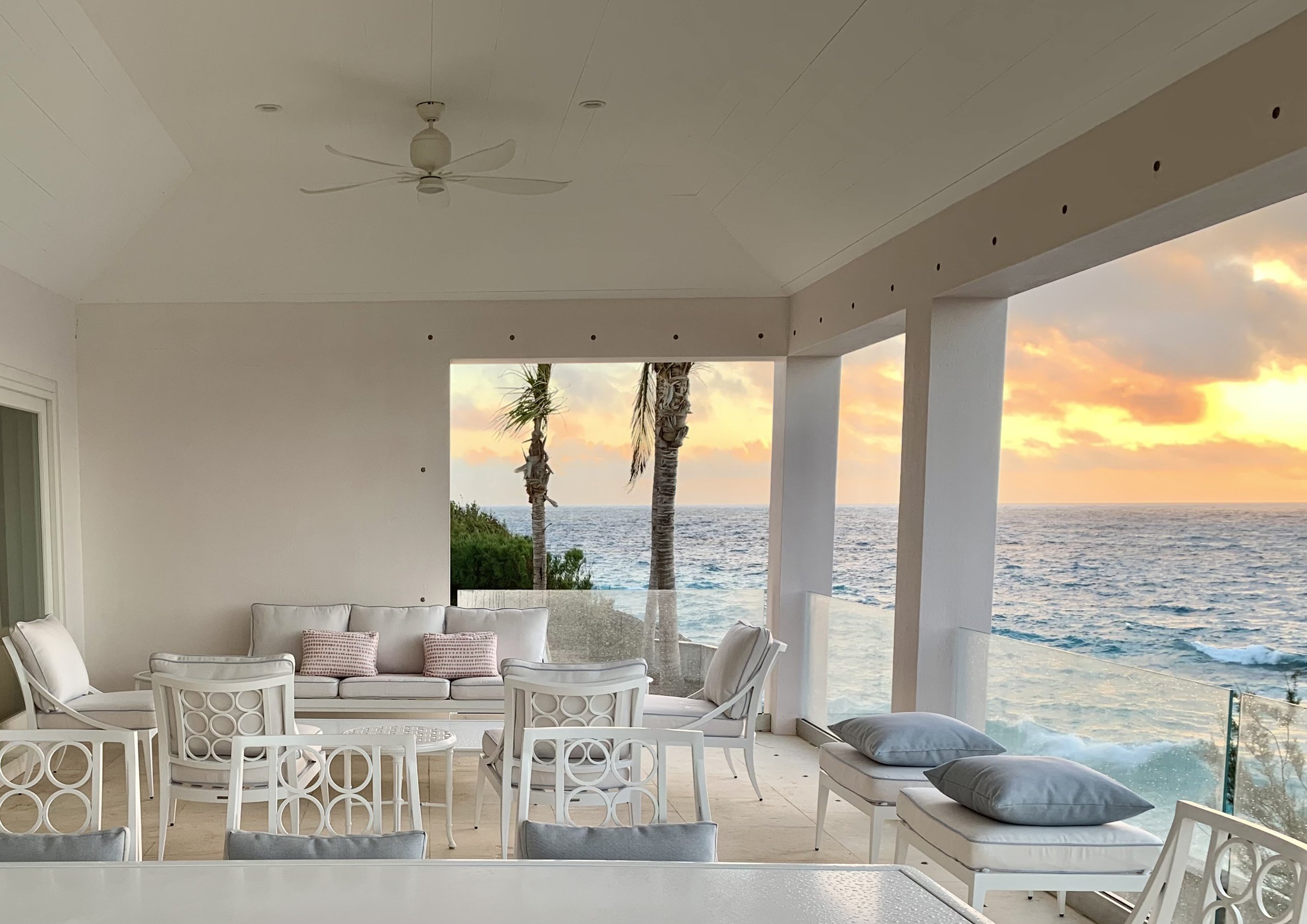Covered oceanfront terrace at sunset, white seating and palms above the water