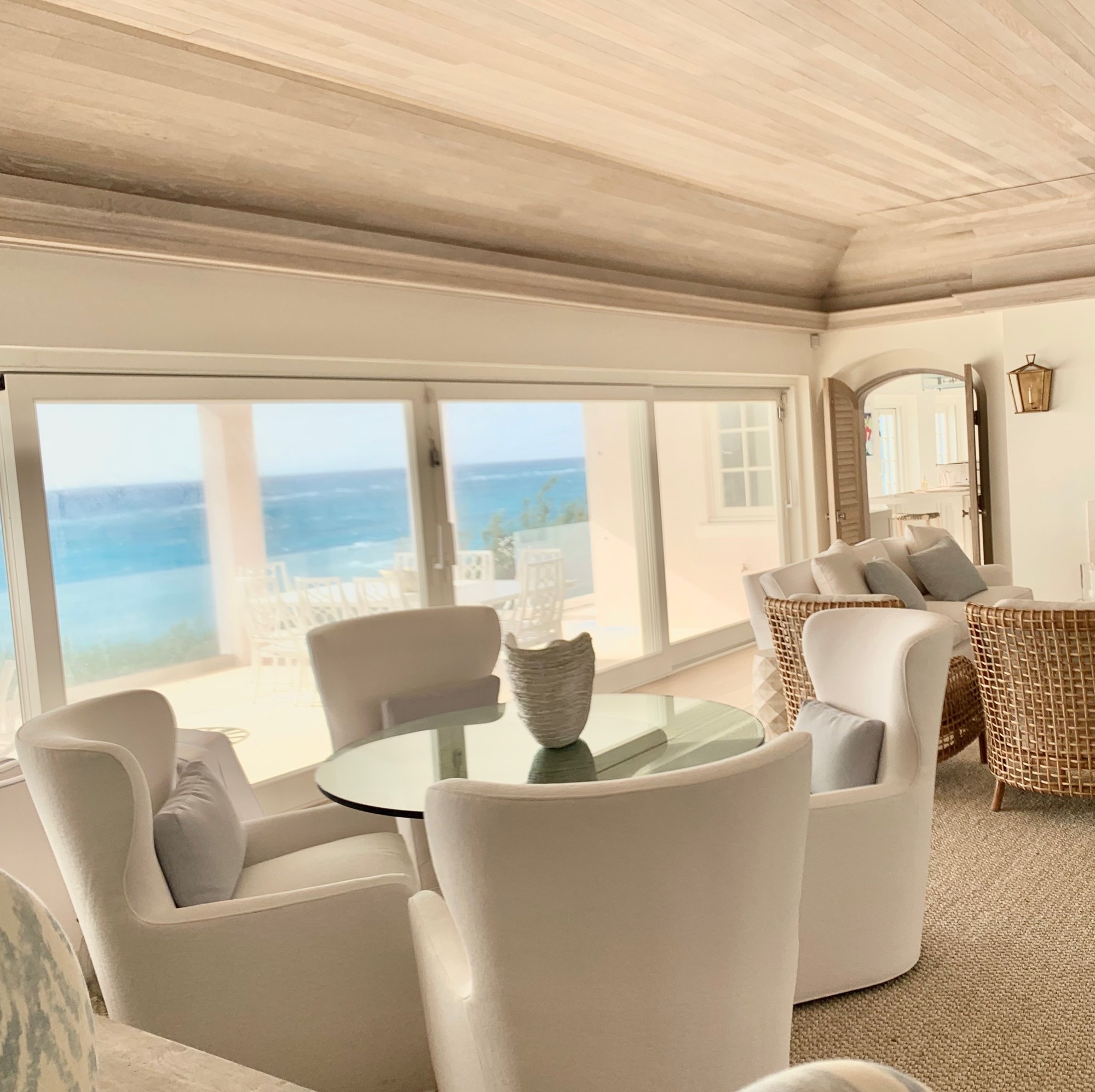 Sitting room of white swivel chairs and wicker facing a wall of glass and the ocean